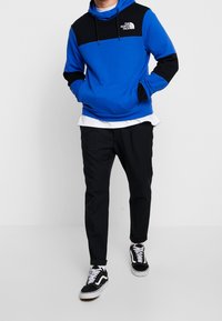 Modrá a černá mikina s kapucí, předním logem, stahovací šňůrkou a klokaní kapsou; v kombinaci s černými kalhotami a klasickými čern�ými teniskami.