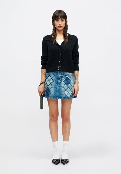 Femme se tenant debout portant un cardigan noir, une chemise blanche, une mini-jupe en denim bleu avec des motifs en losange, des chaussettes blanches, des chaussures noires à bout pointu et tenant une petite pochette.