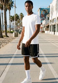 Joven vestido con camiseta blanca, pantalones cortos negros y zapatillas blancas caminando por un paseo marítimo soleado con palmeras y edificios cercanos.