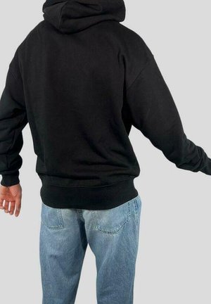 Person iført en sort hættetrøje og lyseblå jeans, set bagfra, med armene let udstrakte til siderne mod en ensfarvet baggrund.
