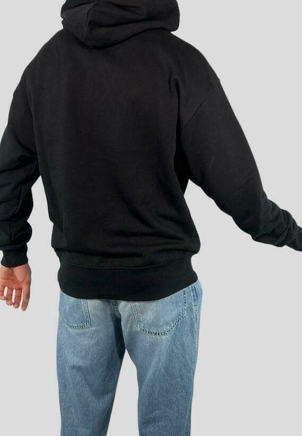 Personne portant un sweat à capuche noir et un jean bleu clair, vue de dos, bras légèrement étendus sur les côtés devant un fond uni.