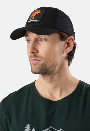 Man draagt een zwarte pet met oranje elandlogo en een donkergroen shirt met wit berg- en boomontwerp, kijkt naar links tegen een lichte achtergrond.