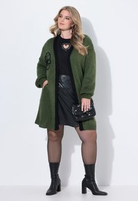 Grüne Strickjacke mit schwarzem Besatz und floraler Stickerei, getragen über einem schwarzen Oberteil und einem Lederrock, kombiniert mit einer schwarzen Handtasche und kniehohen Stiefeln.