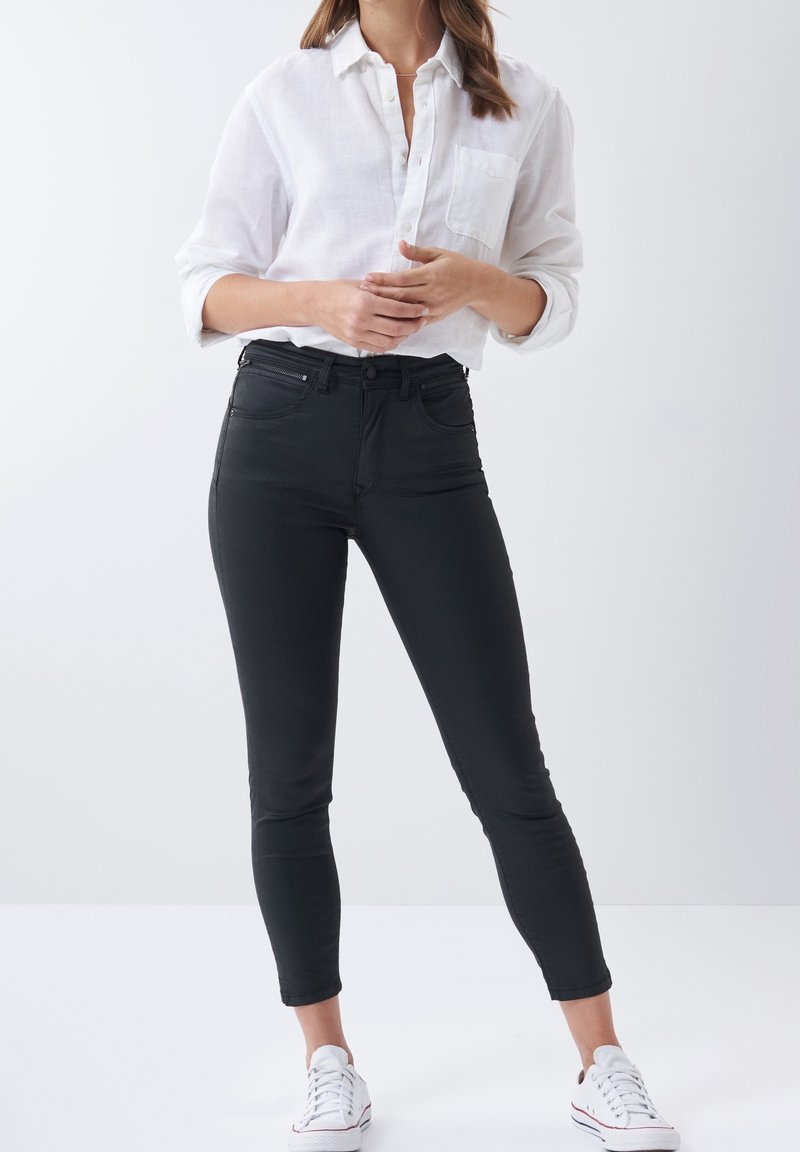 Black skinny jeans with a high waist, featuring front pockets and belt loops. Paired with a white button-up shirt. Worn with white sneakers.