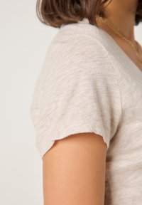 Približen posnetek ramena in zgornjega dela roke osebe, ki nosi svetlo bež kratko majico in zlato verižico.