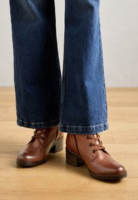 Botas de cuero marrón hasta el tobillo con punta redondeada, detalles de cordones y tacón apilado, combinadas con jeans de mezclilla oscura de pierna acampanada.