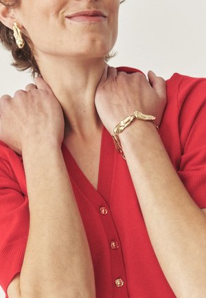 Gioielli in tono oro con un design strutturato, abbinati a un cardigan rosso con bottoni decorativi, che mostrano le braccia incrociate attorno al collo.