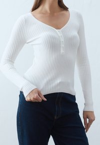 Biała, prążkowana bluzka z długim rękawem i dekoltem w serek, z zapięciem na guziki przypominające perły, połączona z ciemnoniebieskimi dżinsami.