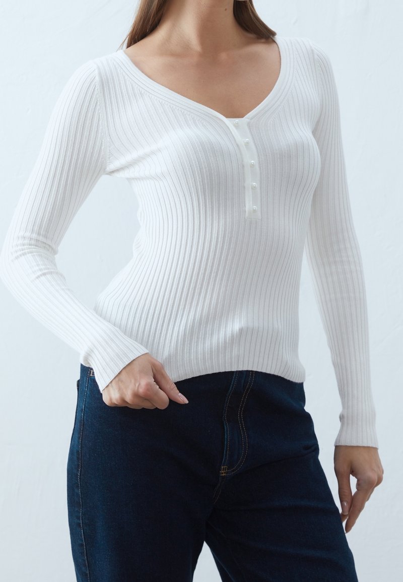 Biała, prążkowana bluzka z długim rękawem i dekoltem w serek, z zapięciem na guziki przypominające perły, połączona z ciemnoniebieskimi dżinsami.