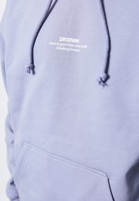 Lila hoodie med dragsko och framficka. Har vit text "DRDENIM" och logotyp. Mjuk tyg med slät struktur.