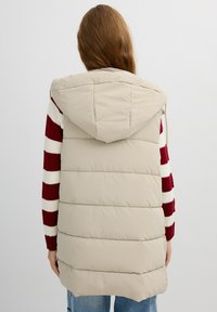Chaleco de plumas beige con capucha, que presenta un diseño acolchado y un cuello alto, llevado sobre un suéter a rayas rojas y blancas. Corte casual.