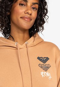 Beige Hoodie mit zwei Herzmotiven, eines schwarz und eines mit Holzmaserung, darunter das Wort "RONY" in Weiß. Texturierter Stoff und Kordelzüge.