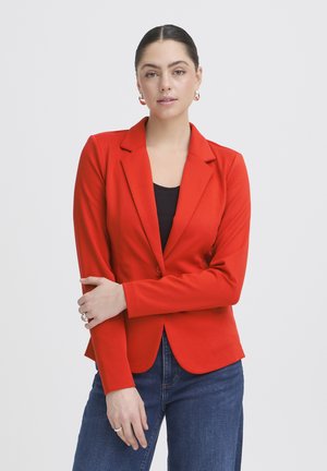 Kvinde iført en tætsiddende rød blazer over en sort top med blå jeans, stående med armene krydset og et neutralt udtryk mod en ensfarvet baggrund.