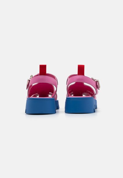 Bakgrunnsvinkel av sandaler med rosa syntetiske stropper, en rød pull-tab, og en tykk blå gummisåle. Spennehelning for justerbar passform.