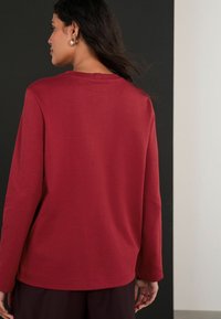 Piros, hosszú ujjú felső, kerek nyakkivágással, puha tapintással és laza szabással. Egyenes alja és minimális varrási részletek jellemzik.