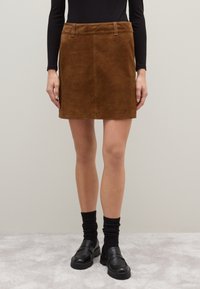 Minifalda de ante marrón con pasadores, combinada con una blusa negra de manga larga, calcetines negros y mocasines negros sobre un fondo de moqueta clara.