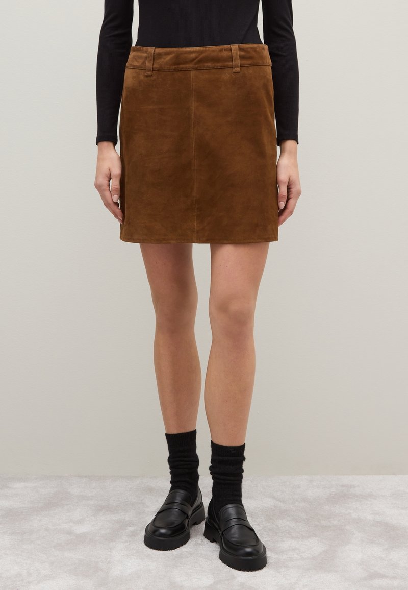 Minifalda de ante marrón con pasadores, combinada con una blusa negra de manga larga, calcetines negros y mocasines negros sobre un fondo de moqueta clara.