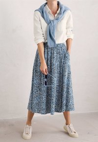 Sininen kukallinen midi-hame, jossa on taskut, yhdistetty valkoiseen napitettuun paitaan ja vaaleansiniseen neuleeseen, joka on solmittu hartioiden ympärille. Valkoiset tennarit käsissä.