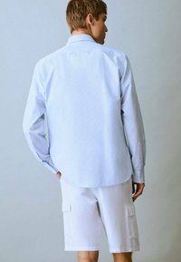 Svetlo modra pajama s dolgimi rokavi s črtami v kombinaciji z belimi kratkimi hlačami s štirimi žepi. Srajca ima ovratnik na gumb in manšete.