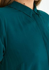 Tealblus med krage, featuring korta ärmar och en slät textur. Inkluderar söm och diskret fickdetalj.