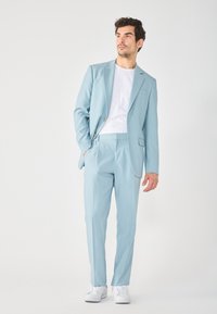 Completo azzurro chiaro con giacca aderente e pantaloni, realizzato in tessuto liscio. T-shirt bianca sotto, abbinata a sneakers bianche.
