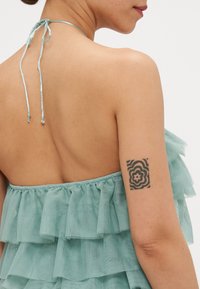 Lyseblå bluse med volanger og halterneck, laget av gjennomsiktig stoff med lagdelt design. Utsatt rygg og blomster tatovering på overarmen.