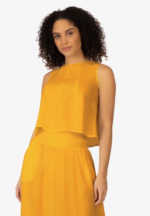 Frau mit lockigem Haar, die ein ärmelloses gelbes Oberteil und einen passenden Rock trägt und vor einem schlichten hellen Hintergrund steht.