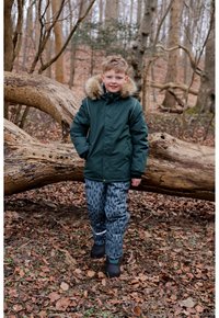 Green insulated jacket with fur-trimmed hood, paired with patterned gray snow pants. Black rubber boots. Outdoor setting with leaves and trees.