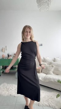 Νεαρή γυναίκα με μαύρο φόρεμα χωρίς μανίκια στέκεται σε ένα φωτεινό σαλόνι με λευκό καναπέ, πράσινη ντουλάπα και μία αφράτη μοκέτα σε πλακάκια στο πάτωμα.
