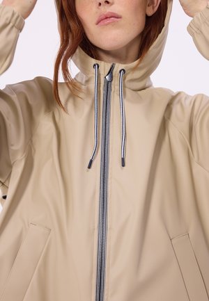 Frau mit roten Haaren, die eine beige Kapuzenjacke mit Frontreißverschluss und gestreiften Kordeln trägt und die Kapuze anpasst.