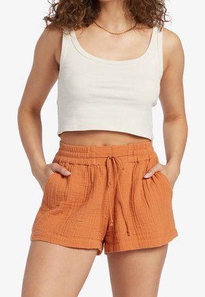 Femme portant un haut court blanc sans manches et un short taille haute orange texturé avec poches, les mains posées à l'intérieur des poches.