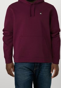 Felpa color bordeaux con tasca frontale e logo Tommy Jeans, indossata con jeans blu, che mostra il torso e le braccia su uno sfondo semplice.