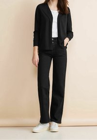 Černý cardigan přes bílý tričko, doplněný o černé džíny s vysokým pasem a bílé tenisky. Hladké textury a ležérní designové detaily.