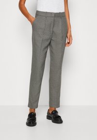 Pantalon gris à motifs avec poches latérales, coupe ajustée et taille ceinturée. Porté avec des mocassins noirs, affichant un style smart-casual.