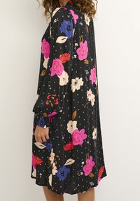 Blommig klänning med svart bakgrund, prydd av rosa, blå och vita blommor. Långa ärmar med elastiska manschetter, avslappnad passform och prickigt mönster.