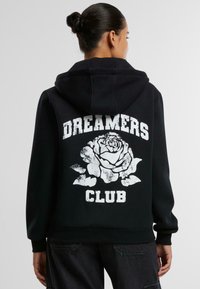 Felpa nera con cappuccio caratterizzata da una grande grafica di una rosa bianca e la scritta "DREAMERS CLUB" sul retro. Tessuto morbido con una vestibilità comoda e cappuccio.