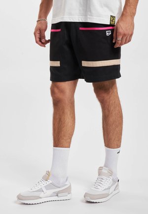 Schwarze Shorts mit pinken und beigen Akzenten, ausgestattet mit Netz-Taschen und einem Logo-Tag. Kombiniert mit weißen Sneakers und gerippten weißen Socken.
