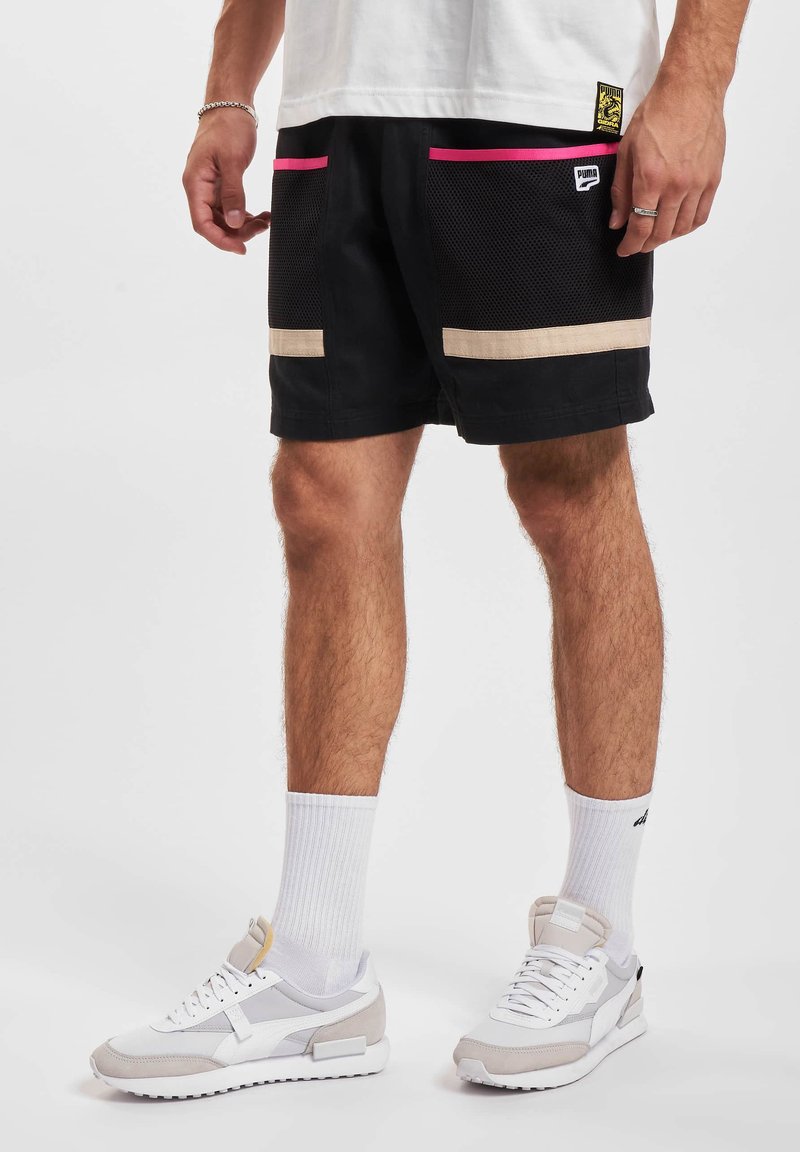 Schwarze Shorts mit pinken und beigen Akzenten, ausgestattet mit Netz-Taschen und einem Logo-Tag. Kombiniert mit weißen Sneakers und gerippten weißen Socken.