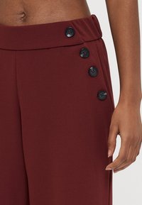 Pantalones de pierna ancha burdeos con una textura suave, que cuentan con una cintura alta y cinco botones negros a lo largo del costado como detalle.