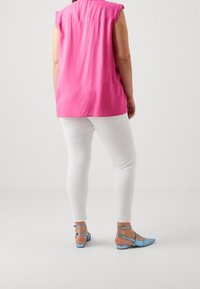 Žena stojící zády, oblečená v jasně růžovém bezsleeve topu, bílých těsných kalhotách a světle modrých nízkých lodičkách s páskem kolem kotníku.