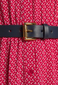 Ceinture en cuir bleu marine avec une boucle dorée, attachée sur une robe rouge ornée de petits motifs floraux blancs et noirs.