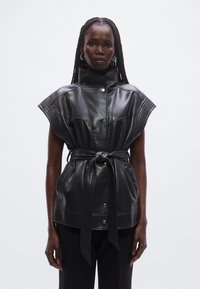 Zwarte leren mouwloze jas met een hoge kraag, riem in de taille en horizontale stikdetails. Voorzien van drukknoopsluiting aan de voorkant en strakke lijnen.