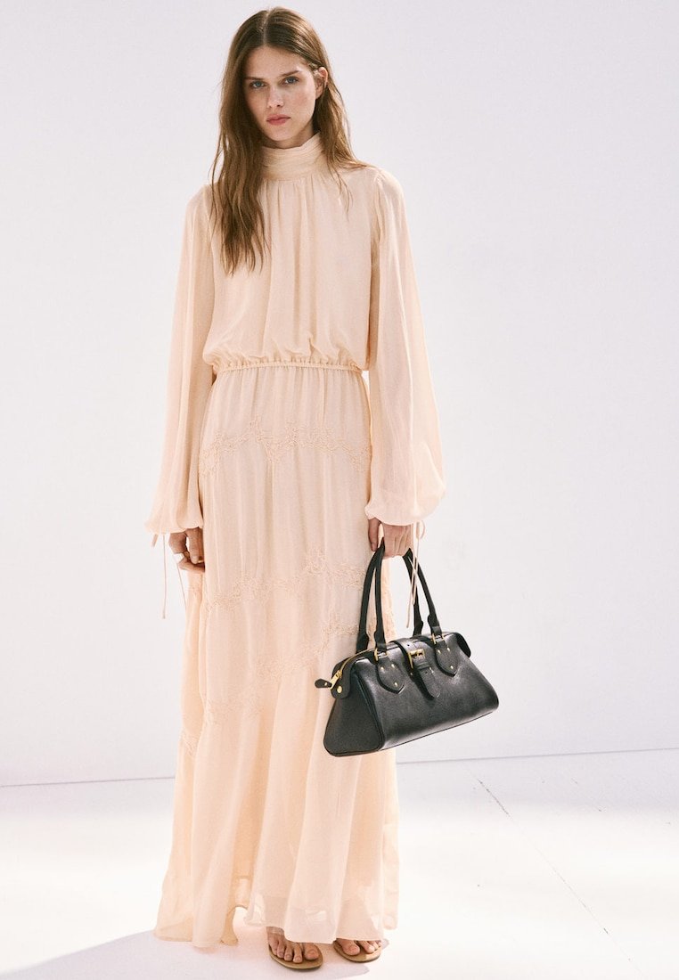 Jeune femme en longue robe beige tenant un sac à main noir, debout devant un fond clair uni.