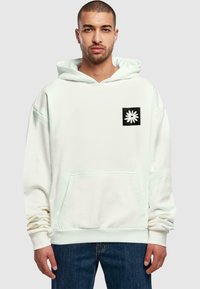 Sudadera blanca de gran tamaño con un bolsillo canguro frontal, que presenta un logotipo cuadrado negro con un diseño de flor blanca en la parte izquierda del pecho.