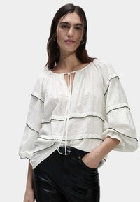 Blusa bianca in tessuto texturizzato, con dettagli neri e un nodo al collo. Caratterizzata da maniche ampie e design a strati. Indossata con pantaloni neri.