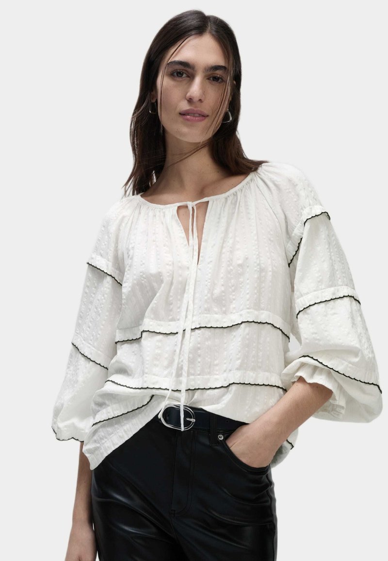 Blusa bianca in tessuto texturizzato, con dettagli neri e un nodo al collo. Caratterizzata da maniche ampie e design a strati. Indossata con pantaloni neri.