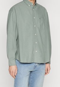 Chemise vert clair à boutons, avec une poche poitrine unique, un col et une fermeture à boutons, fabriquée en tissu doux avec une coupe décontractée.