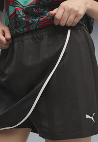 Άτομο που ρυθμίζει μαύρα αθλητικά σορτς Puma με λευκή επένδυση και ελαστική μέση, φορώντας πουκάμισο με πράσινο και κόκκινο μοτίβο.