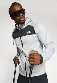 Light grey zip-up hoodie with black accents, featuring a logo on the chest, worn by a person holding trekking poles.