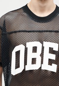 Μαύρο διαπνέον πουκάμισο με μεγάλα λευκά γράμματα "OBE" στο στήθος, φορεμένο από ένα άτομο με ανοιχτόχρωμο δέρμα, που δείχνει τον λαιμό και μερικό πρόσωπο.